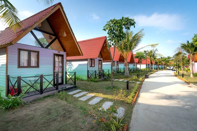 Rivi värikkäitä siirtolapuutarhamökki-mökkejä eloisassa trooppisessa maisemassa rehevän vehreän kasvillisuuden keskellä.