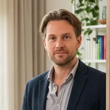 A man standing in a bright living room, showcasing smart home controls on a wooden wall, with large windows and plants.